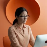Mulher usando óculos e camisa listrada laranja, concentrada enquanto trabalha em um computador de mesa em um ambiente com decoração em tons de laranja.
